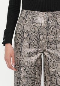 Κοντινό πλάνο ενός ατόμου που φοράει παντελόνι ψηλής μέσης με γκρι και μαύρο σχέδιο φιδιού και μαύρη ριμπ μπλούζα μακρυμάνικη με κουμπιά στον καρπό.