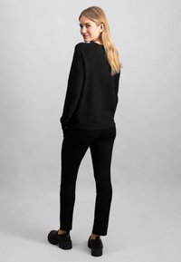 Čierny oversized sveter s dlhými rukávmi, skombinovaný s úzkymi čiernymi nohavicami. Model má na sebe mohutné čierne topánky a pózuje zozadu.