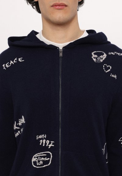 Haut de survêtement zippé bleu marine, présentant des designs brodés en blanc comprenant du texte et des symboles. Texture douce, avec une poche avant et une capuche ajustable.
