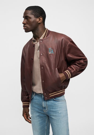 Καφέ δερμάτινο μπουφάν τύπου μπόμβερ με ριμπ ύφασμα στη λαιμόκοψη και τις μανσέτες, με μπεζ ρίγες και κεντημένο λογότυπο στο στήθος.