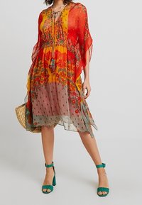Vestito fluido e multicolore con motivi floreali in rosso, arancione e verde. Tessuto trasparente, lunghezza al ginocchio, con scollo a legatura e dettagli con frange.