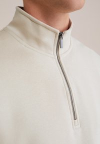 Pullover a mezza zip beige chiaro con collo alto, realizzato in tessuto morbido. Caratterizzato da una cerniera argentata e una texture liscia.