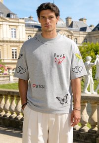 Jeune homme portant un t-shirt gris oversize avec des griffonnages colorés et du texte, debout à l'extérieur près d'une architecture européenne classique.