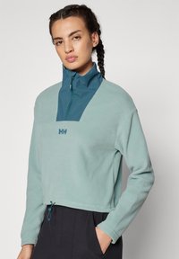 Sudadera de forro polar verde claro con cuello alto azul, cremallera frontal y logo. Presenta un diseño corto y cordón ajustable en el dobladillo.