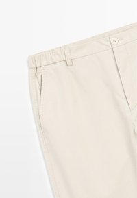 Pantalon en coton beige avec fermeture par bouton, taille élastique, poches latérales et passants de ceinture, montré de la partie supérieure de la jambe jusqu'à la zone de la taille.