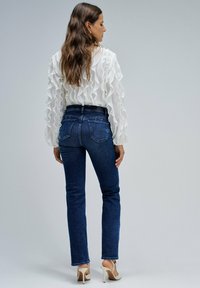 Camisa blanca con textura arrugada, combinada con jeans de pierna recta en azul oscuro. El atuendo incluye un cinturón negro y sandalias de tacón alto de color claro.
