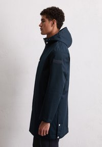 Man in marineblauwe capuchonjas die zijwaarts tegen een effen lichte muur staat, hand ontspannen naast zich, jas met knoopdetails en een mouwpatch.