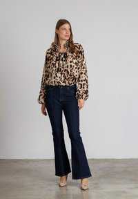 Blusa de estampado de leopardo con un cuello alto y detalle de lazo, combinada con jeans acampanados oscuros. Zapatos beige con acento de lazo completan el look.