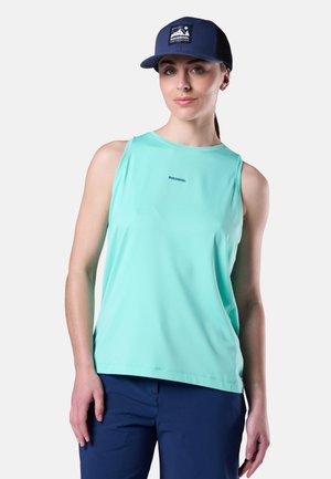 Kobieta w jasnoniebieskim, bezrękawnym topie z logo "Rossignol", granatowych spodniach i granatowej czapce Rossignol, stojąca na jednolitym tle.