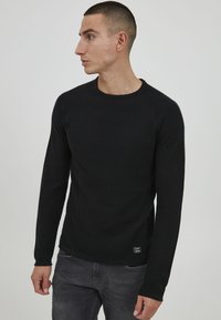 Zwarte gebreide trui met lange mouwen, een textuurpatroon, ronde halslijn en raglanmouwen. Heeft een klein logo-label aan de onderzoom.