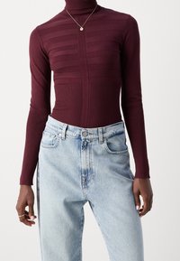 Haut col roulé bourgogne avec une texture côtelée horizontale et un design ajusté, associé à un jean taille haute légèrement usé de couleur bleu clair.