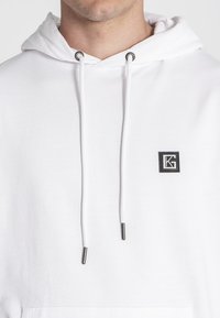 Sudadera blanca de tela suave con capucha ajustable, que presenta un pequeño logotipo negro en el lado izquierdo del pecho y acentos de metal plateado.