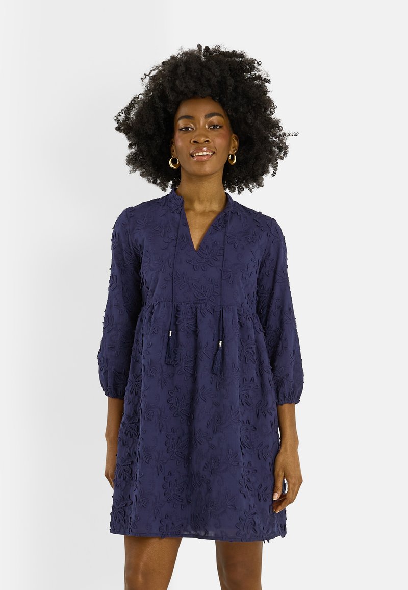 Femme aux cheveux bouclés naturels portant une robe texturée à fleurs bleu marine à manches trois-quarts et des boucles d'oreilles anneaux dorées, debout devant un fond blanc.