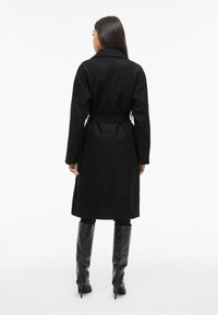 Kvinne står vendt bortover iført en beltestengt svart frakk, svarte leggings og svarte høye hæler, mot en enkel hvit bakgrunn.