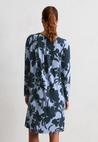 Donna con i capelli rossi raccolti in una coda di cavallo, che indossa un abito azzurro chiaro a grandi fantasie floreali scure, in piedi contro un muro bianco semplice.