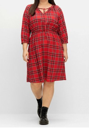Kobieta w czerwonej sukience w kratę do kolan z rękawami o długości do łokcia i czarnych, sznurowanych botkach idąca do przodu na białym tle.