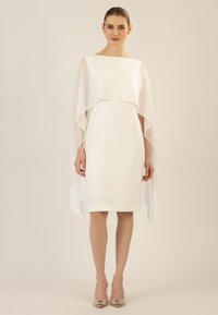 White dress with a layered sheer cape, knee-length, structured design, and a round neckline. Worn with embellished beige heels.
