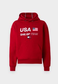 Rød hættetrøje med frontlomme, der har "USA," det amerikanske flag, "SAILGP TEAM," og Tommy Hilfiger-logoet på brystet.
