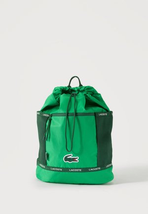 Sac à dos à cordon vert Lacoste avec poche zippée à l'avant, poches latérales en mesh et écussons logo sur le devant et les bretelles.