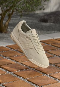 Beige volnočasové tenisky se šněrováním a texturovanou podrážkou, vznášející se nad dřevěnými dlaždicemi a štěrkem venku poblíž rostliny.