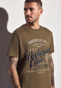 Muž s krátkými vlasy a tetováním na pažích, oblečený ve tmavě zeleném tričku "Workwear Crew", hledící do strany na jednobarevném pozadí.