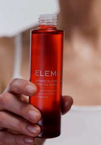 Rød glassflaske med kroppsolje og hvit skrukork. Merket har "ELEMIS" og produktdetaljer i hvit tekst. Glatt overflate.