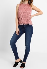 Krajkový top v barvě prachové růže s květinovým vzorem a vroubkovaným lemem, ve spojení s tmavými úzkými džínami a černými mules s otevřenou špičkou.