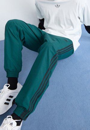 Osoba siedzi na niebieskiej podłodze, ubrana w zielone spodnie dresowe z czarnymi paskami, białe buty Adidas, czarne skarpetki i białą koszulkę Adidas.
