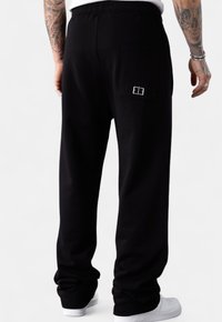 Man wearing black sweatpants with a back pocket and white embroidered logo, paired with white sneakers on a plain background.