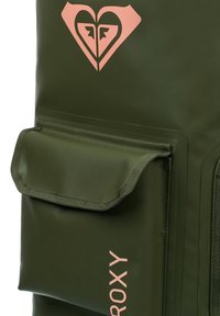 Sac à dos étanche vert avec une poche frontale à rabat, présentant un logo rose et une surface texturée. Comprend une fermeture éclair et des coutures renforcées.