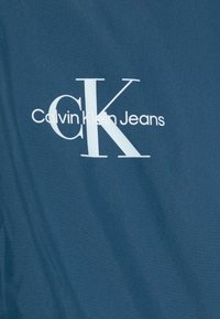 Calvin Klein Jeans-logoen på et mørkeblått stoff, med hvite bokstaver der "CK" er tydelig vist og "Calvin Klein Jeans" nedenfor.