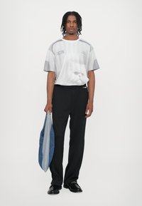 Hvid grafisk t-shirt med et kattemotiv, grå accenter, parret med sorte bukser. Modellen holder en blå denimtaske og har sorte sko på.