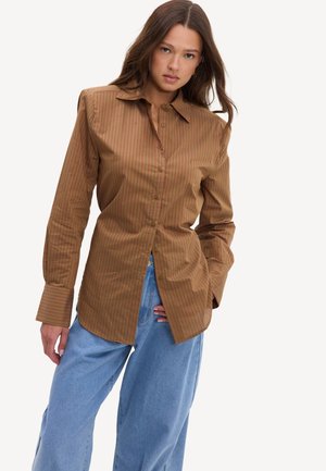 Jeune femme portant une chemise à rayures marron avec boutons et un jean bleu clair ample, debout, une main dans la poche sur fond blanc.
