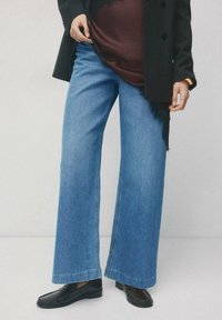 Spodnie typu wide-leg z wysokim stanem, wykonane z niebieskiego denimu, z czystym wykończeniem i minimalnym przeszyciem. Połączone z czarnym płaszczem i ciemnym obuwiem.