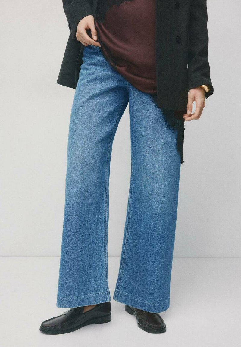 Spodnie typu wide-leg z wysokim stanem, wykonane z niebieskiego denimu, z czystym wykończeniem i minimalnym przeszyciem. Połączone z czarnym płaszczem i ciemnym obuwiem.