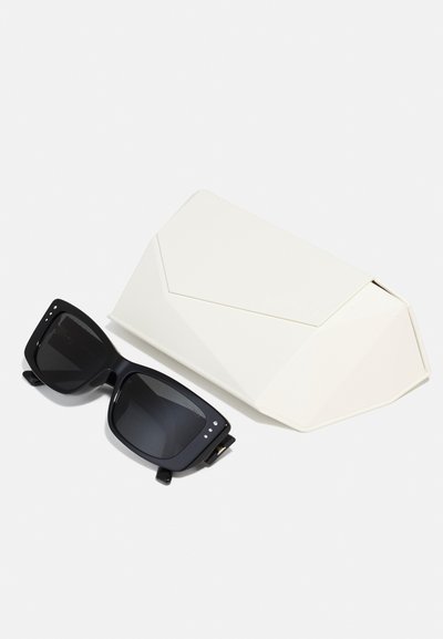 Ochelari de soare negri cu ramă pătrată și lentile întunecate, însoțiți de un etui geometric alb, având textură netedă și un design minimalist.