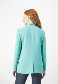 Blazer turchese con maniche lunghe, rever a punta e uno spacco sul retro. Tessuto liscio con dettagli minimi e un logo discreto sul colletto.