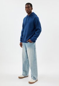 Sudadera azul de tejido suave con un bolsillo tipo canguro, combinada con unos vaqueros holgados de un azul claro y zapatillas crema con suelas marrones.