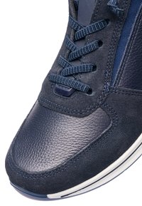 Detailní záběr špičky tenisky tmavě modré barvy s texturovanou kůží, semišovými panely, sladěnými tkaničkami a bílým okrajem gumové podrážky.