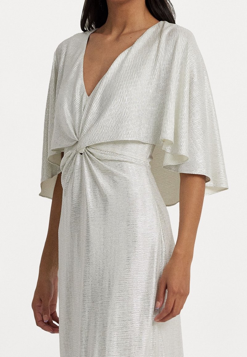 Kvinna klädd i en silver metallisk klänning med djup V-ringning, vriden knutdetalj vid midjan och breda, flödande korta ärmar.