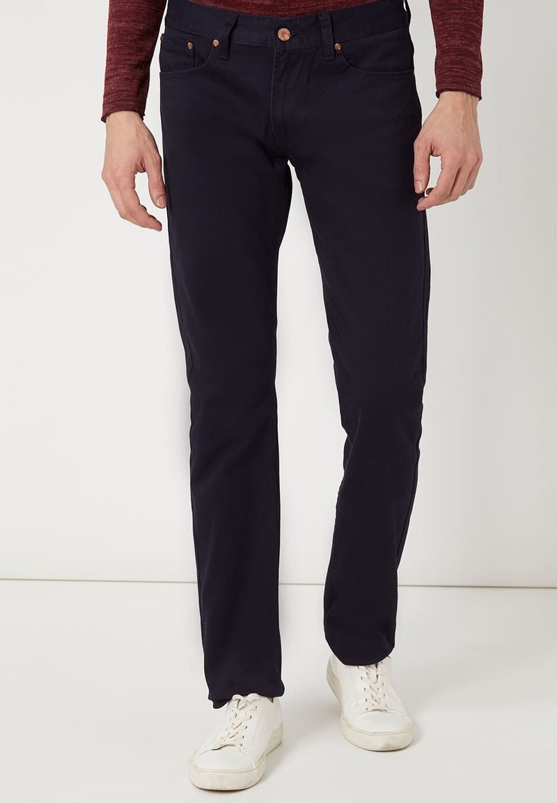 Vaqueros azul marino de corte slim, fabricados en denim. Presentan cinco bolsillos, detalles de botones de cobre y una textura suave. Combinados con zapatillas deportivas blancas.
