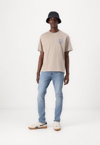 Beige bomull T-skjorte med blå grafisk detalj, kombinert med lyseblå denimjeans og kremfargede sneakers med svarte og brune aksenter.
