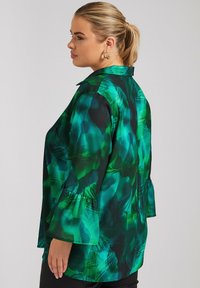 Blusa verde con un estampado de hojas tropicales, de corte holgado, con cuello y mangas acampanadas. Textura suave, colores vibrantes y dobladillo curvo.