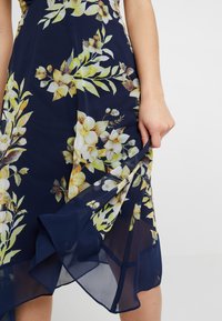 Bloemenjurk met een marineblauwe achtergrond, gele en witte bloemen en groene bladeren; lichtgewicht stof met een gelaagde, vloeiende zoom.