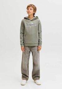 Zelena kapuca s belim grafičkim motivom planine i logotipom brenda, s prednjim džepom, nošena s svijetlosivim širokim trapericama i bijelim tenisicama.