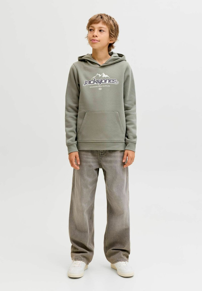 Zelena kapuca s belim grafičkim motivom planine i logotipom brenda, s prednjim džepom, nošena s svijetlosivim širokim trapericama i bijelim tenisicama.