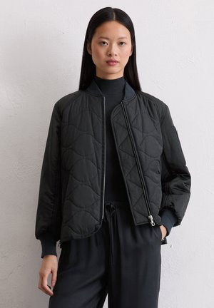 Schwarze gesteppten Bomberjacke mit Reißverschluss, geripptem Kragen und Bündchen. Verfügt über eine Seitentasche und eine glatte Textur. Trägt sie über einem schwarzen Rollkragenpullover.