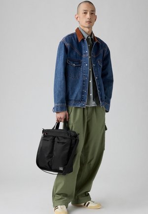Svart nylon tote-bag med to frontlommer, båret av en person i en denimjakke med brun krage og grønne cargo-bukser.