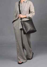 Bolso de hombro de cuero negro con un diseño plano, acentos en hardware dorado y una correa ancha, llevado por una persona vestida con un atuendo de lino gris claro.