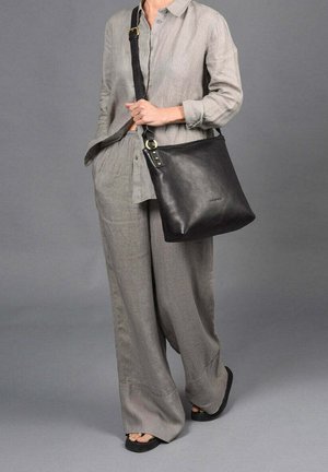 Black leather shoulder bag with a flat design, gold hardware accents, and a wide strap, carried by a person in light gray linen attire.